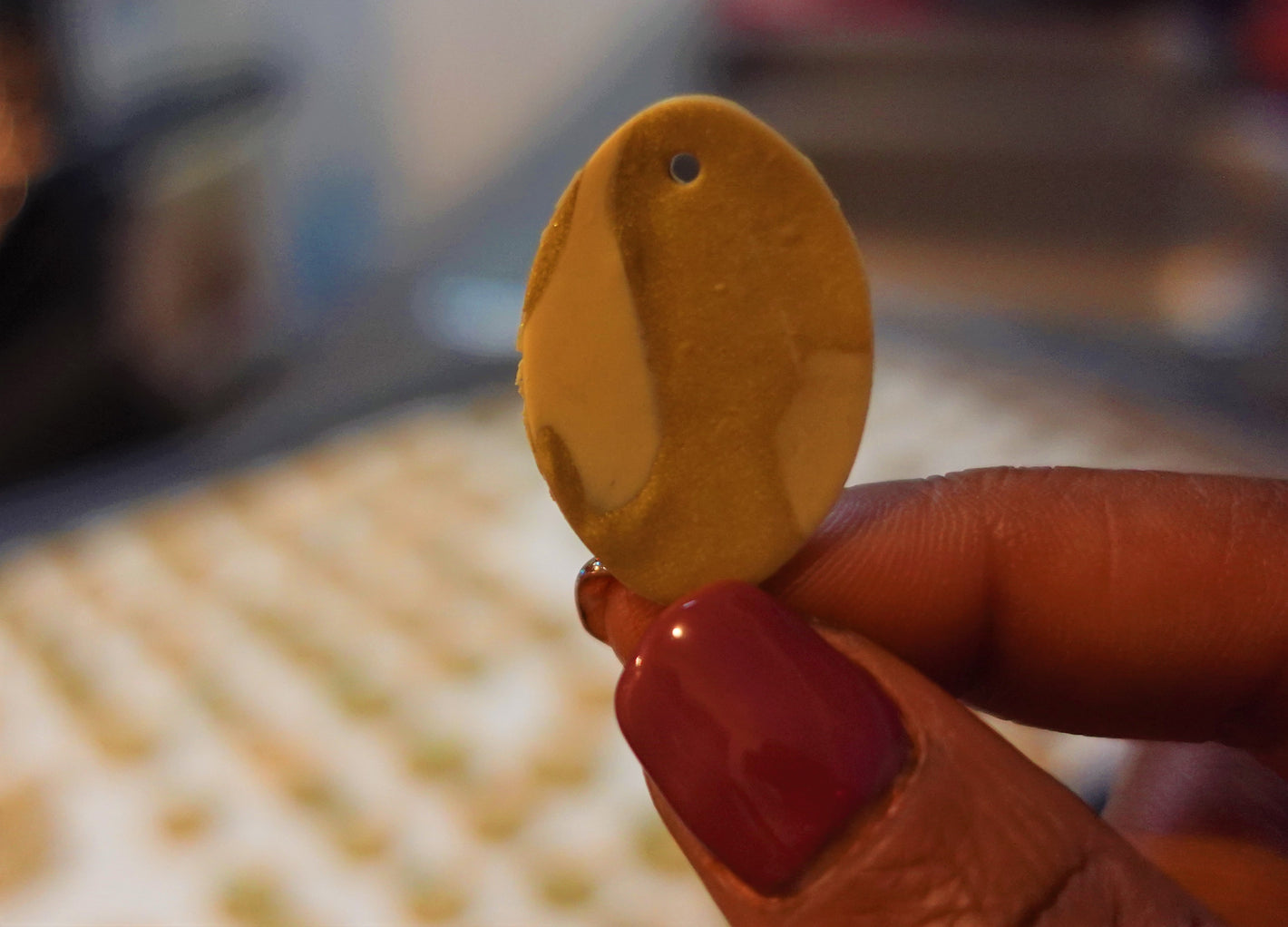 close up of fingers with purple nail polish holding beige and gold oval polymer clay earring piece with baking sheet lined with additional beige pieces blurred in the background 